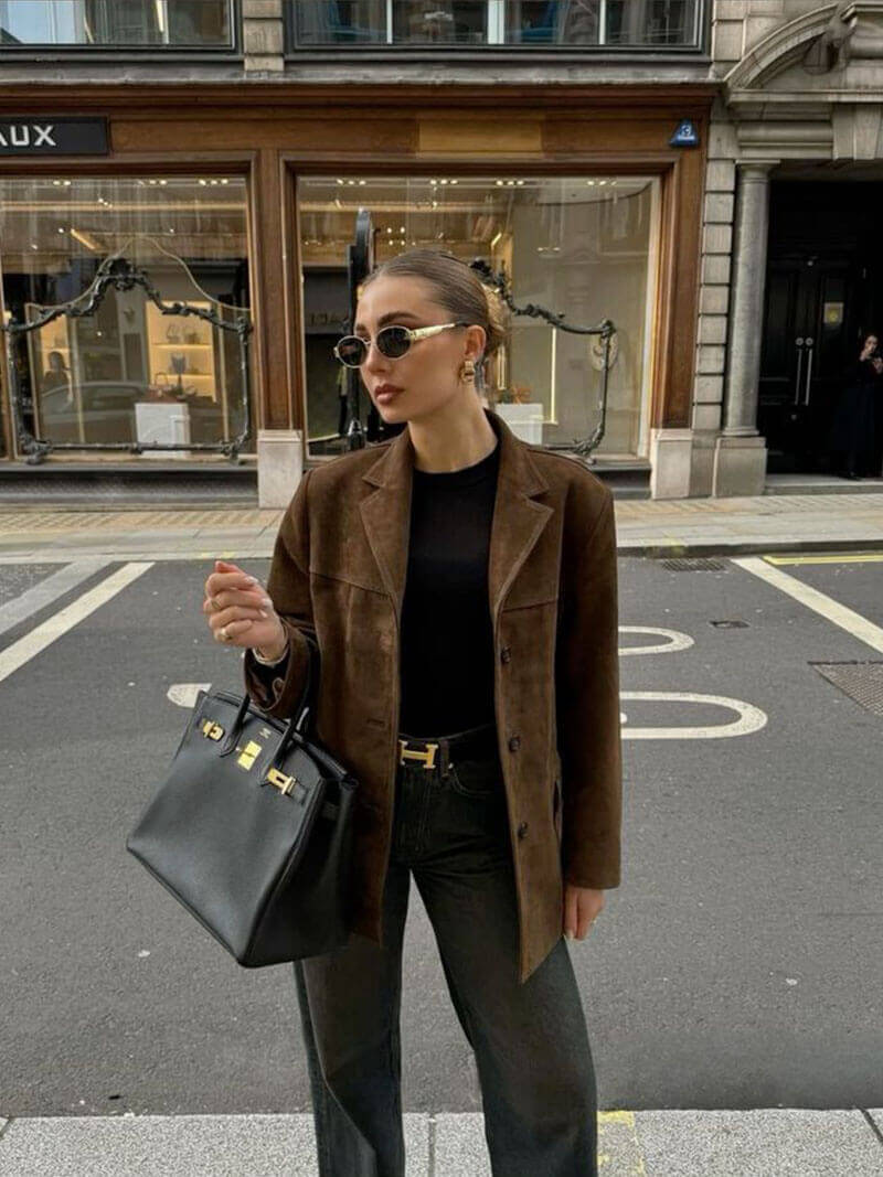 Woman wearing a brown suede leather jacket, black top, sunglasses, and holding a black handbag, standing on a city street.