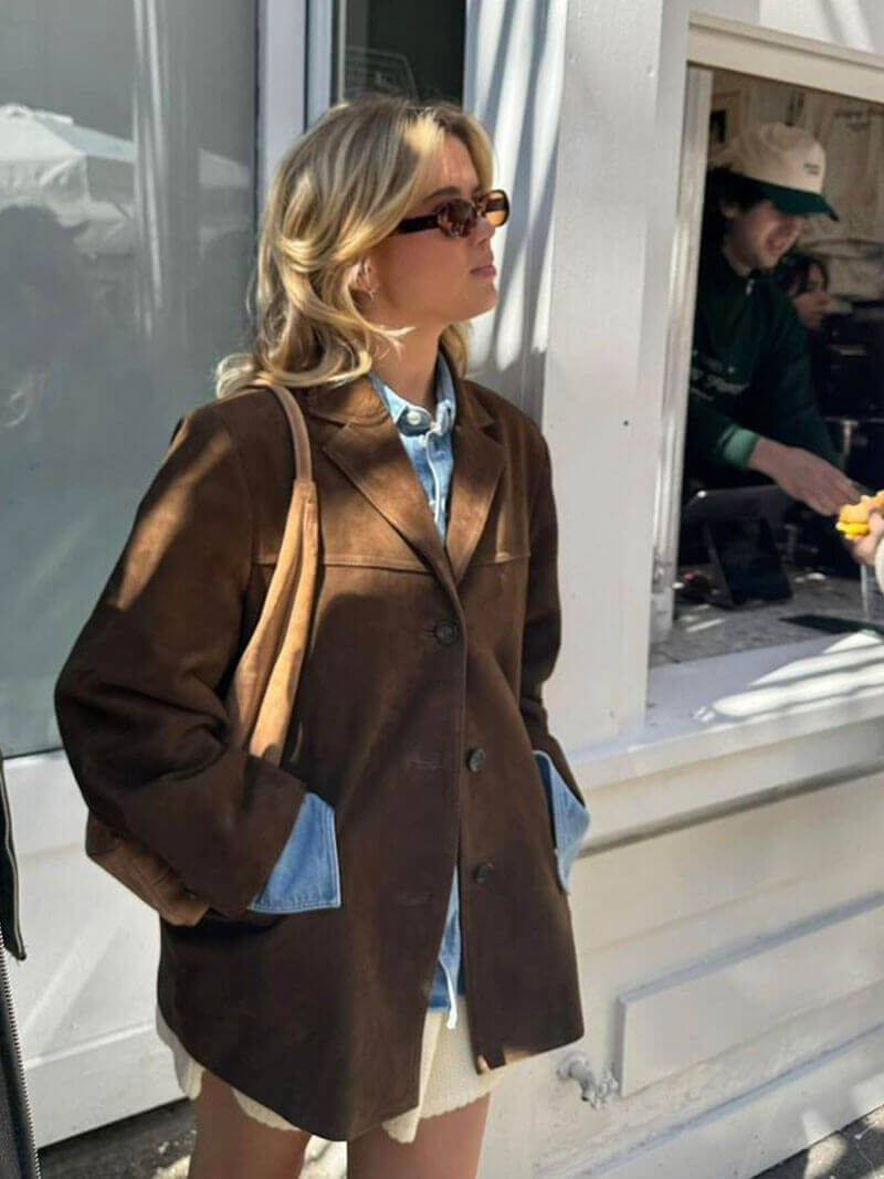 Woman wearing a stylish brown suede leather jacket over a blue shirt and sunglasses, standing casually outdoors.