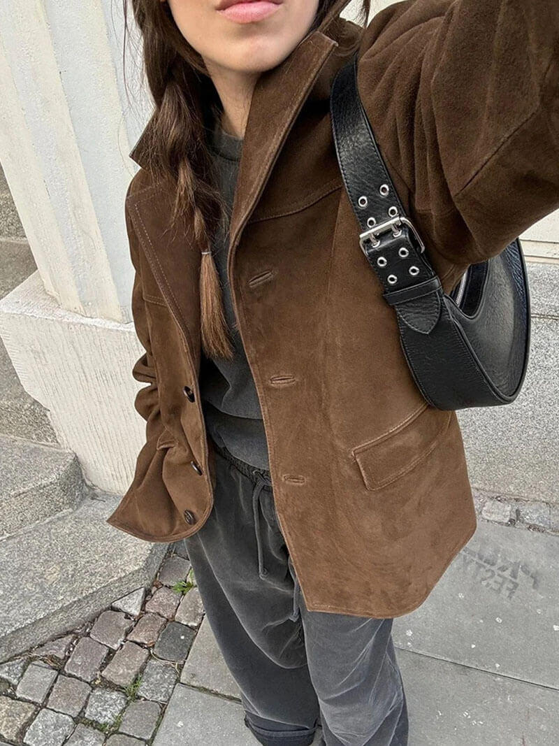 Woman wearing a brown suede leather jacket with a black shoulder bag, standing outdoors on a cobblestone street.