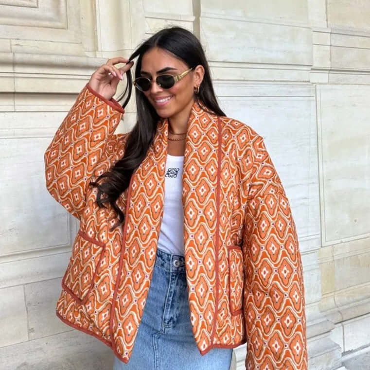 Woman wearing stylish orange patterned Swannie Jacket with sunglasses and denim skirt, smiling while standing against a stone wall.