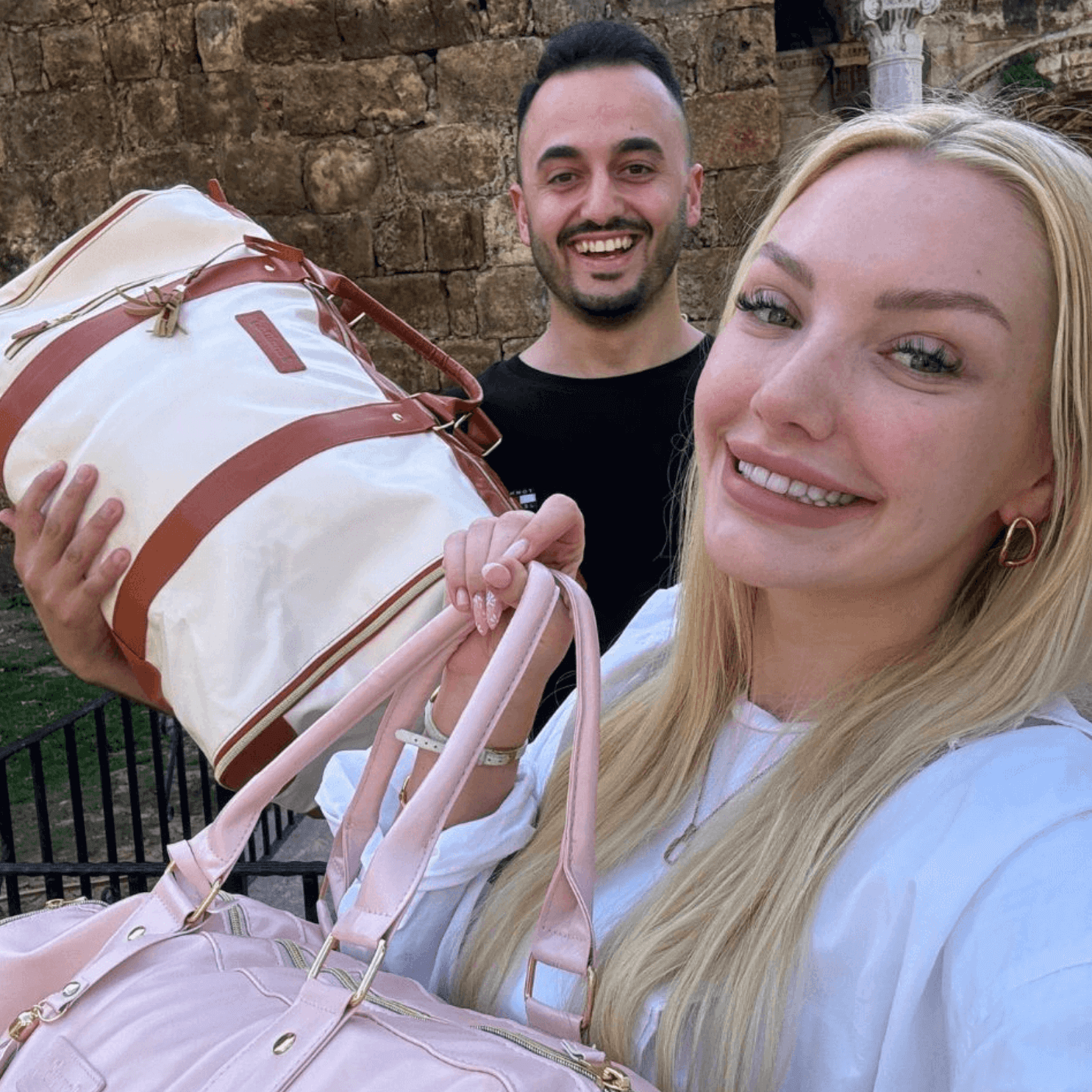 Two people smiling and holding stylish travel bags outdoors.